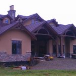 A large brown house featuring a spacious porch, surrounded by greenery and sunlight.